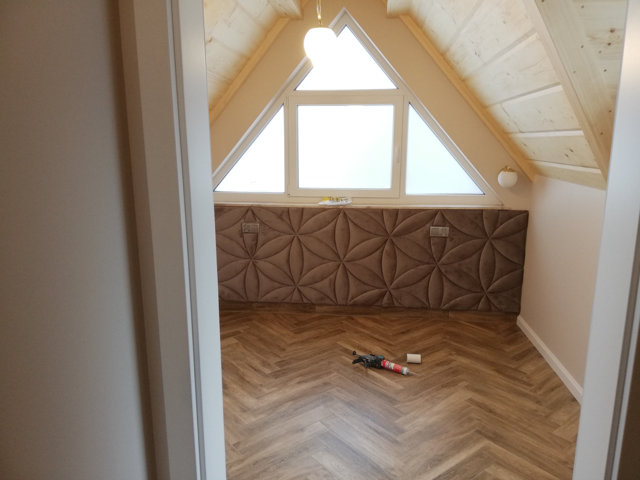 Poddasze z oknem trójkątnym, drewnianą boazerią, panelami ściennymi w kolorze brązu i parkietem ułożonym we wzór jodełki; na podłodze leży pistolet do silikonu i tuba z silikonem.