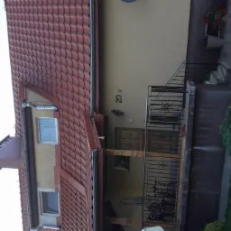 Dom jednorodzinny z brązową dachówką, widoczne rynny, numer domu 202, wejście z metalową balustradą i fragmentem ogrodu.