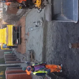 Przygotowanie podłoża pod kostkę brukową: ubijanie piasku zagęszczarką wibracyjną, widoczny wykop, ciężarówka z piaskiem w tle, pracownik w odzieży roboczej nadzorujący prace.