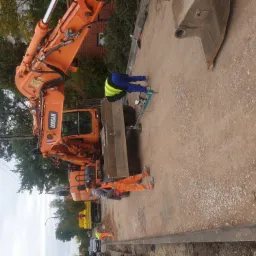 Pomarańczowa koparka Doosan na placu budowy, pracownicy w odblaskowych kamizelkach i kombinezonach, jeden z nich używa specjalistycznego urządzenia pomiarowego do gruntu.