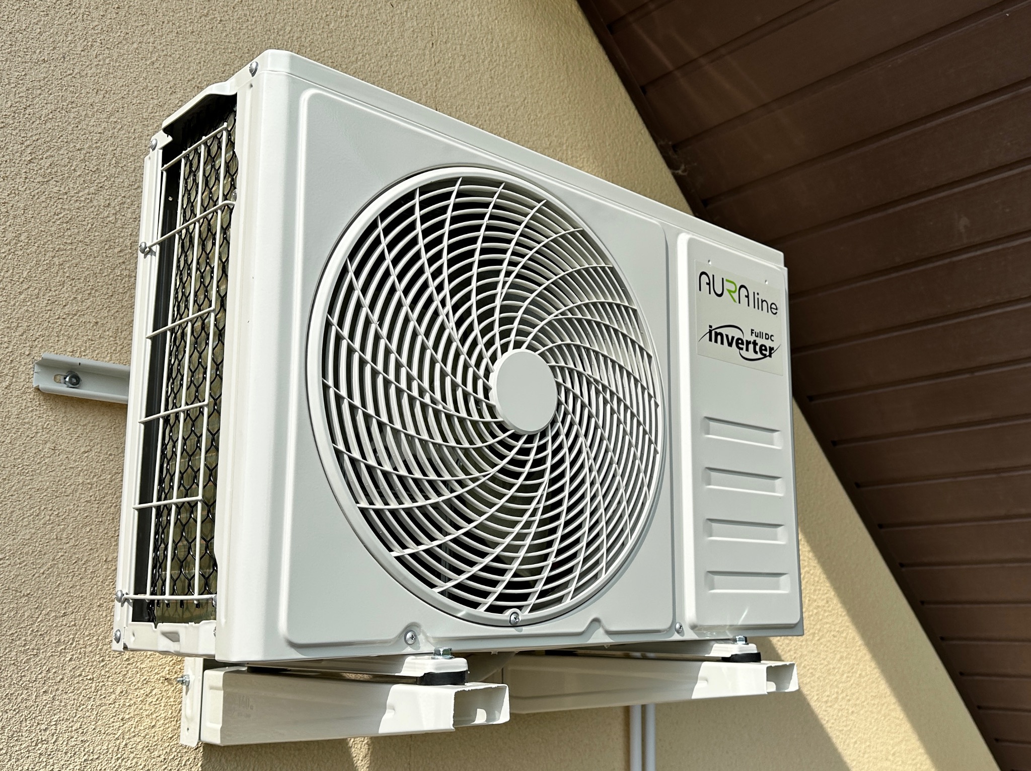 Biała jednostka zewnętrzna klimatyzacji Aura Line Inverter zamontowana na elewacji budynku, widoczna kratka wentylacyjna i elementy montażowe.