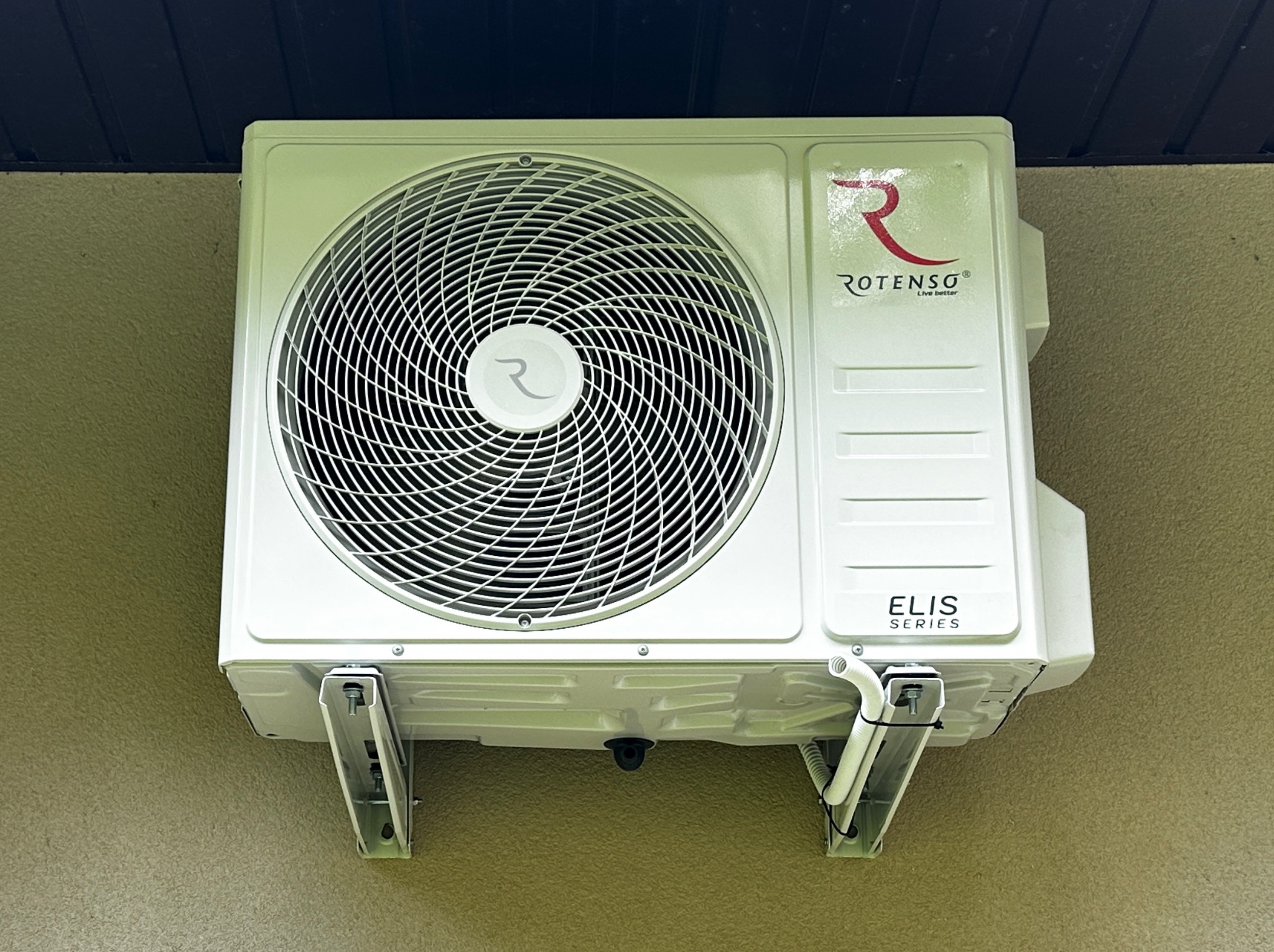 Biały klimatyzator Rotenso ELIS Series zamontowany na ścianie budynku, widoczne wsporniki i przewody. Urządzenie z bliska, z logo producenta i charakterystyczną spiralą wentylatora.