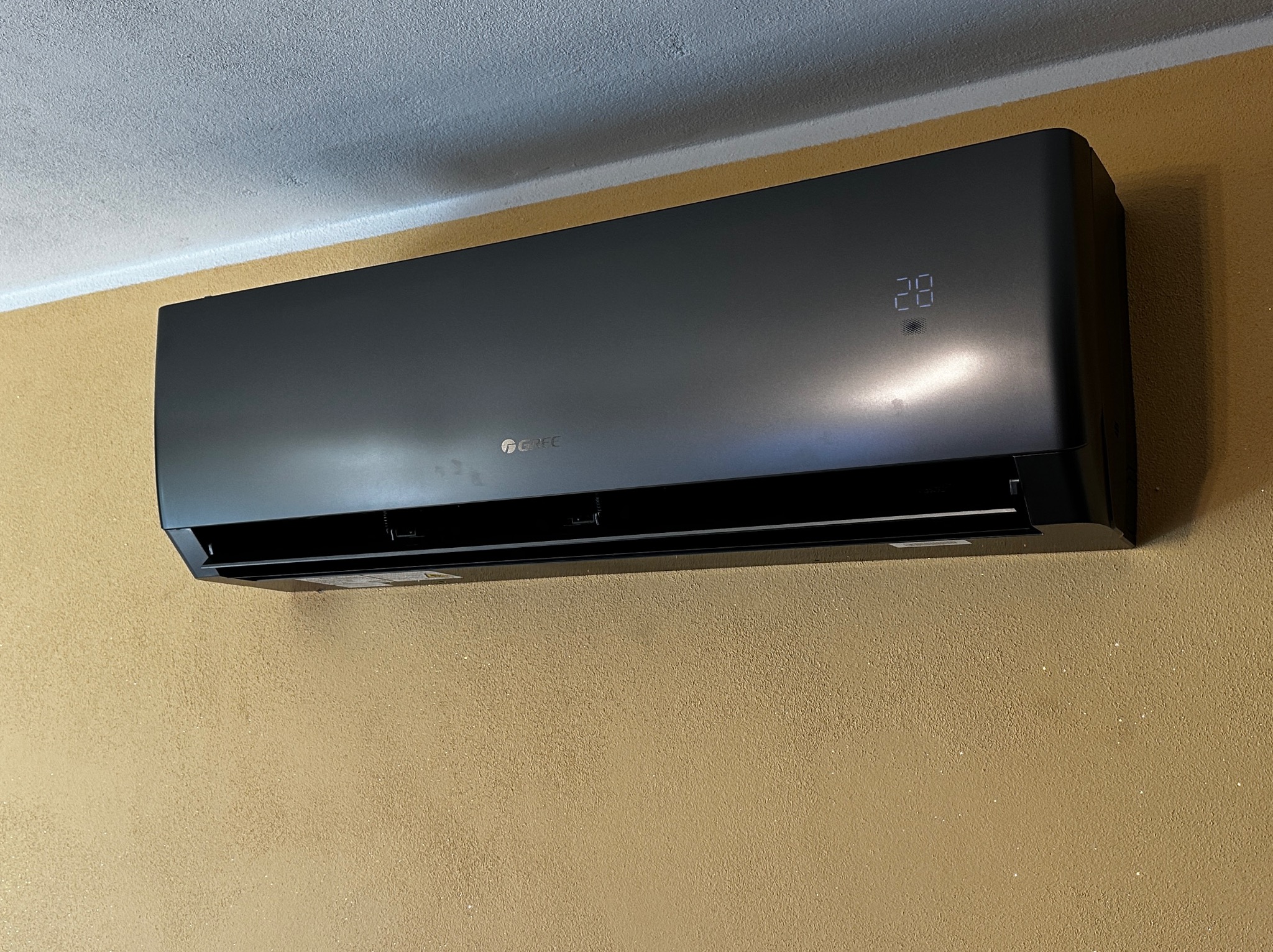 Czarny klimatyzator Gree zamontowany na ścianie o fakturze baranka. Na wyświetlaczu widoczna temperatura 28 stopni. Urządzenie zawieszone blisko sufitu.