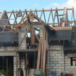 Budowa domu murowanego w trakcie realizacji: ściany z bloczków, drewniana konstrukcja dachu z deskowaniem, widoczne materiały budowlane i częściowo wzniesiony komin.