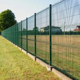 Długi, zielony, panelowy płot z podmurówką w Górze, oddzielający posesję od drogi, z widocznymi domami w tle.