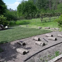 Rozkładanie trawy z rolki w ogrodzie. Widoczne fragmenty ułożonej trawy, rolki trawy oraz narzędzia do układania na przygotowanym podłożu, a w tle zieleń drzew i krzewów.