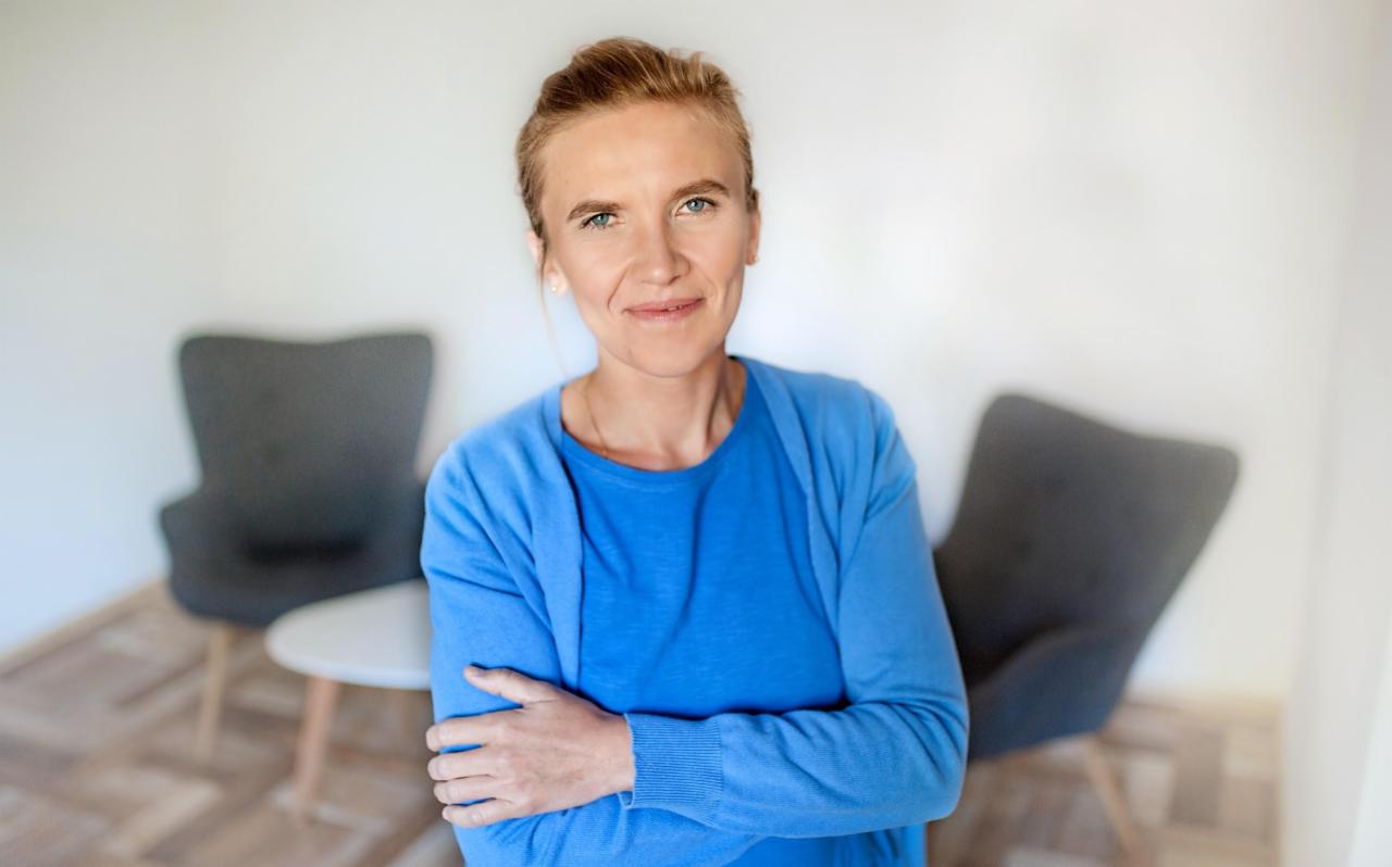 Portret uśmiechniętej kobiety w niebieskim swetrze, z założonymi rękami na tle jasnego pomieszczenia z dwoma szarymi fotelami i małym stolikiem.