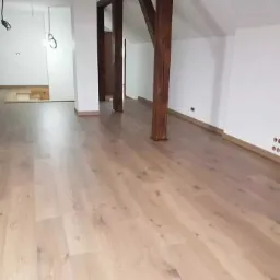 Jasne, drewniane deski podłogowe ułożone w przestronnym wnętrzu z białymi ścianami i drewnianymi słupami konstrukcyjnymi. Fragment podłogi w głębi pomieszczenia jeszcze nie wykończony.