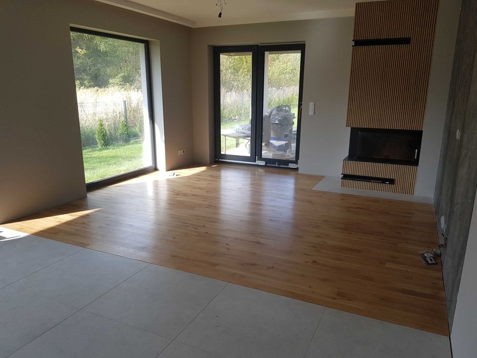 Wnętrze w trakcie wykańczania: połączenie ułożonej drewnianej podłogi z szarymi płytkami, widoczny fragment ściany z kominkiem obudowanym drewnianymi lamelami, duże okno z widokiem na zieleń.