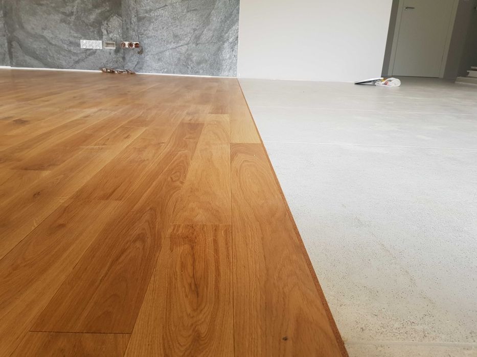 Połowa podłogi wyłożona jasnym parkietem, druga połowa surowy beton, w tle szara ściana imitująca kamień i biała ściana z drzwiami.