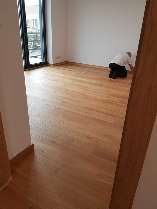Jasny, dębowy parkiet ułożony w pustym pokoju, z widocznym mężczyzną docinającym listwę przypodłogową przy ścianie w rogu pomieszczenia, przez otwarte drzwi widać fragment podłogi i ściany.