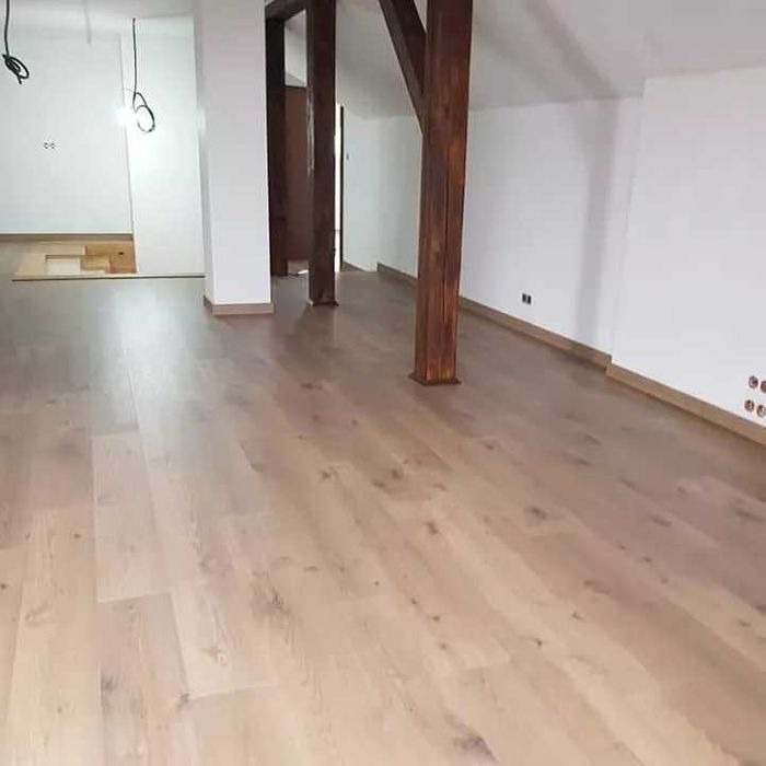 Jasne, drewniane deski podłogowe ułożone w przestronnym wnętrzu z białymi ścianami i drewnianymi słupami konstrukcyjnymi. Fragment podłogi w głębi pomieszczenia jeszcze nie wykończony.