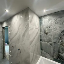 Montaż płytek imitujących marmur w łazience, widoczne krzyżyki dystansowe, fragment wanny i instalacja wodna przed armaturą.