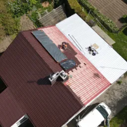 Widok z drona na remontowany dach domu: część dachu pomalowana na biało, część z brązową dachówką, pracownik zabezpiecza fragment dachu plandeką, obok stoi biały bus.