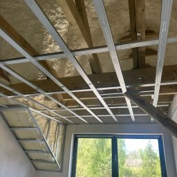 KAR-BUD - Poddasze ocieplone pianą, z widoczną konstrukcją sufitu podwieszanego z profili. W tle okno z widokiem na zieleń. Prace wykończeniowe w toku.