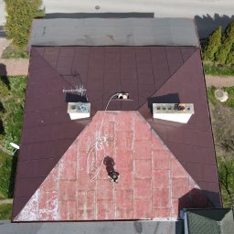 KAR-BUD - Widok z góry na pracownika na dachu domu z brązową blachą, z widocznymi kominami, anteną i elementami otoczenia, takimi jak drzewa i chodnik.