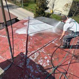 KAR-BUD - Czyszczenie dachu z farby za pomocą myjki ciśnieniowej. Widoczny pracownik w trakcie pracy, z częściowo oczyszczonym dachem w kolorze czerwonym i szarym.