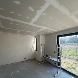 KAR-BUD - Wnętrze w trakcie prac wykończeniowych: sufit i ściany z płyt gipsowo-kartonowych, widoczne łączenia i szpachlowanie, drabina z narzędziami w pomieszczeniu z oknem.