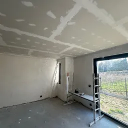 Wnętrze w trakcie prac wykończeniowych: sufit i ściany z płyt gipsowo-kartonowych, widoczne łączenia i szpachlowanie, drabina z narzędziami w pomieszczeniu z oknem.