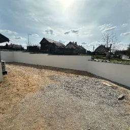 Świeżo pomalowany, biały mur oporowy w ogrodzie, z widocznymi domami w tle i błękitnym niebem z chmurami. Teren przygotowany pod dalsze prace.