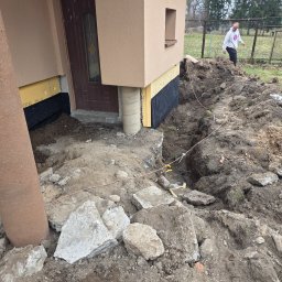 KAR-BUD - Odsłonięty fundament domu z izolacją pionową i drenażem. Wykop wokół budynku, widoczne elementy instalacji elektrycznej. Osoba w tle.