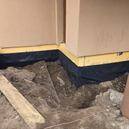 KAR-BUD - Ocieplenie fundamentów budynku z użyciem żółtego styropianu i czarnej folii kubełkowej, widoczne wykopane fundamenty i fragment ściany budynku w beżowym kolorze.