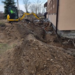 KAR-BUD - Żółta koparka JCB przy wykopie fundamentów budynku. Pracownik przy ścianie budynku. Stosy ziemi i gruzu. Prace ziemne w toku, przygotowanie do dalszych etapów budowy.