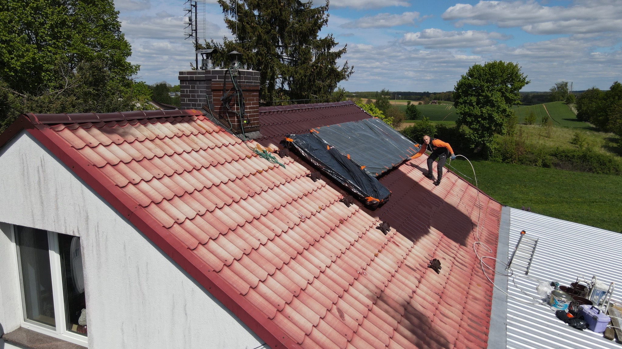 Praca na dachu: mężczyzna w pomarańczowej bluzie zabezpiecza czerwoną dachówkę plandeką. Widoczne narzędzia, drabina i krajobraz w tle.