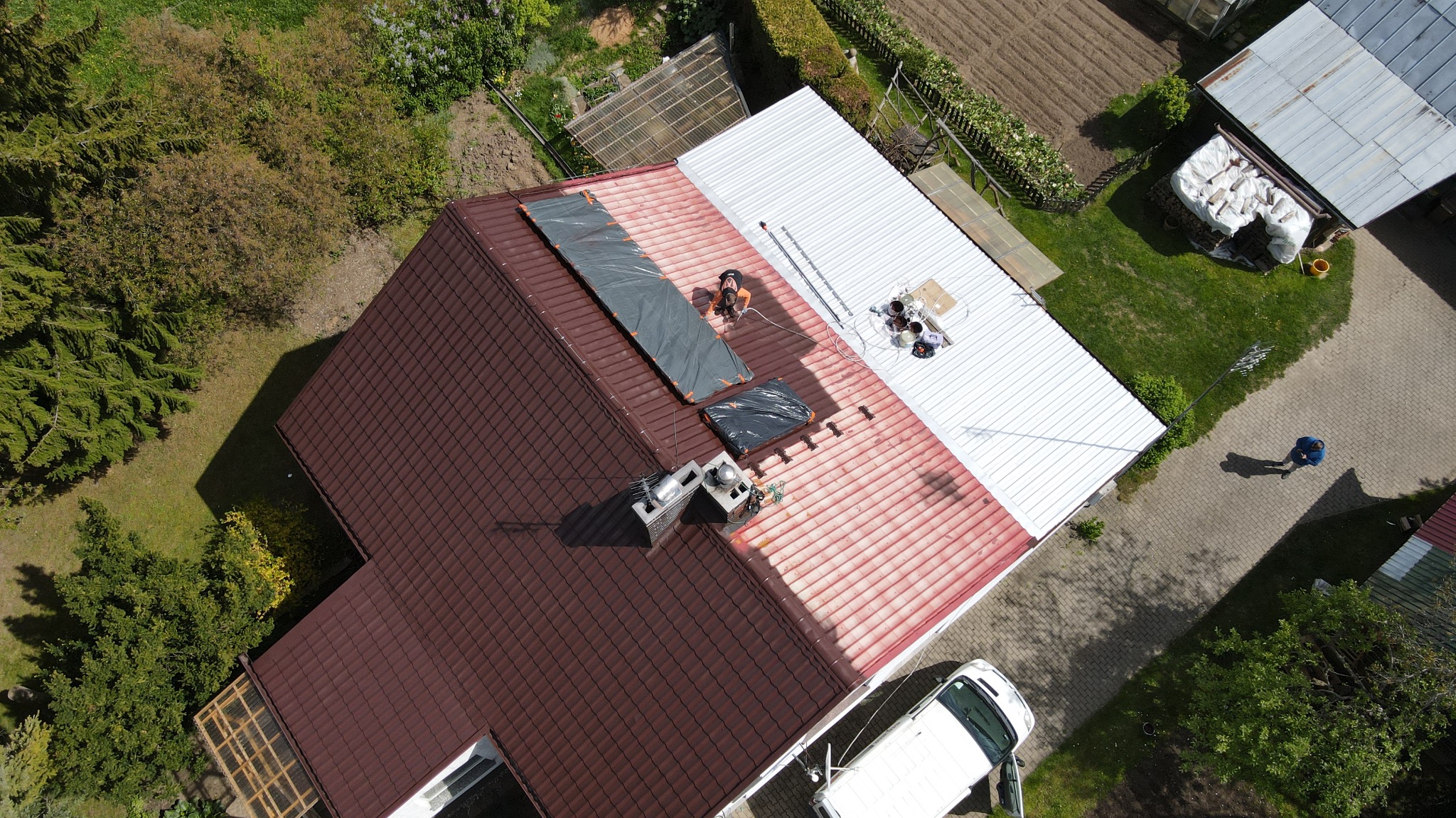 Widok z drona na remontowany dach domu: część dachu pomalowana na biało, część z brązową dachówką, pracownik zabezpiecza fragment dachu plandeką, obok stoi biały bus.