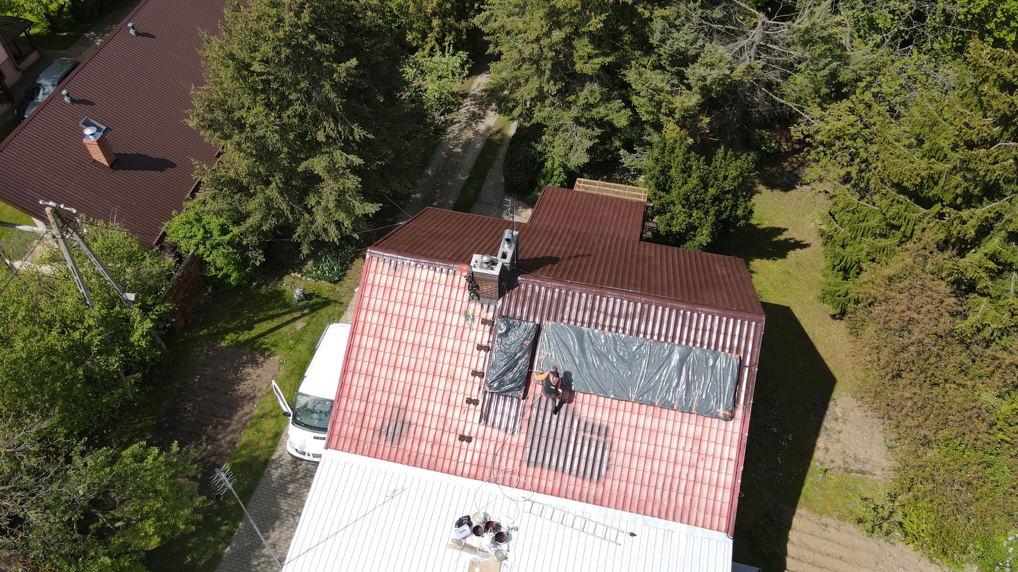 Prace na dachu z blachodachówki z lotu ptaka: malowanie fragmentu dachu na czerwono, zabezpieczone obszary. Widoczny pracownik, drabina, puszki z farbą i otoczenie zieleni.