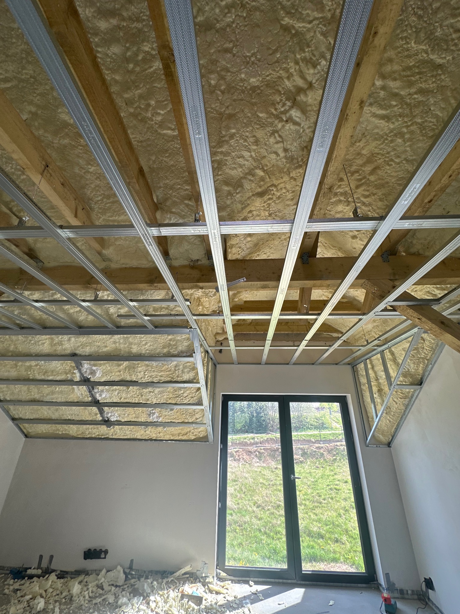 Poddasze z ociepleniem piankowym i metalowym stelażem pod płyty gipsowe, widok od dołu, okno dachowe z widokiem na zieleń, resztki piany na podłodze.