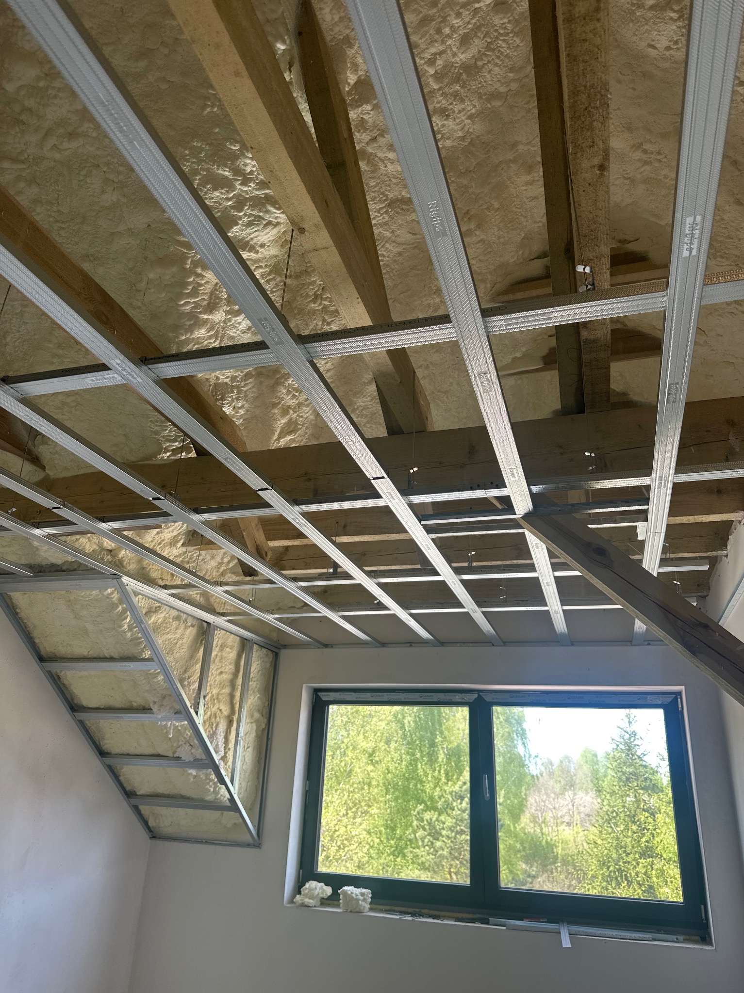 Poddasze ocieplone pianą, z widoczną konstrukcją sufitu podwieszanego z profili. W tle okno z widokiem na zieleń. Prace wykończeniowe w toku.