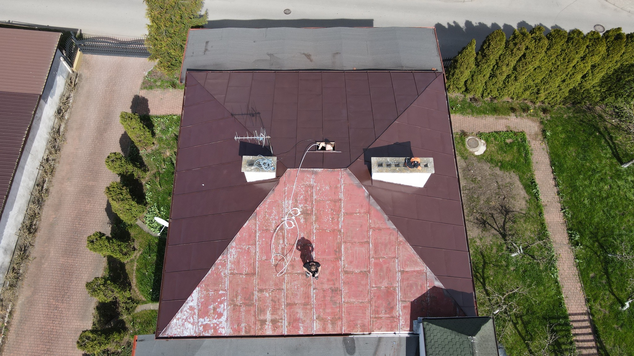 Widok z góry na pracownika na dachu domu z brązową blachą, z widocznymi kominami, anteną i elementami otoczenia, takimi jak drzewa i chodnik.