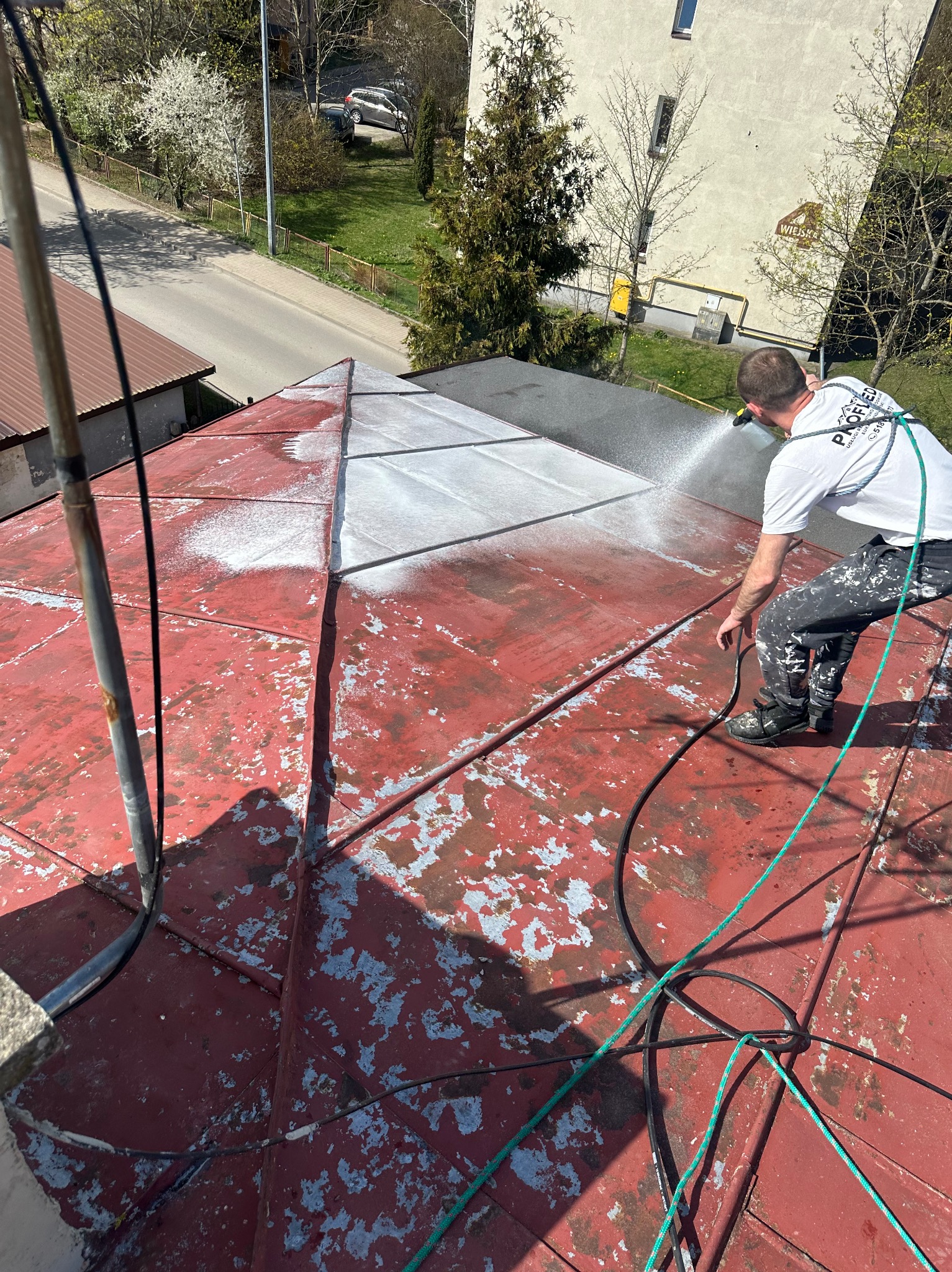 Czyszczenie dachu z farby za pomocą myjki ciśnieniowej. Widoczny pracownik w trakcie pracy, z częściowo oczyszczonym dachem w kolorze czerwonym i szarym.