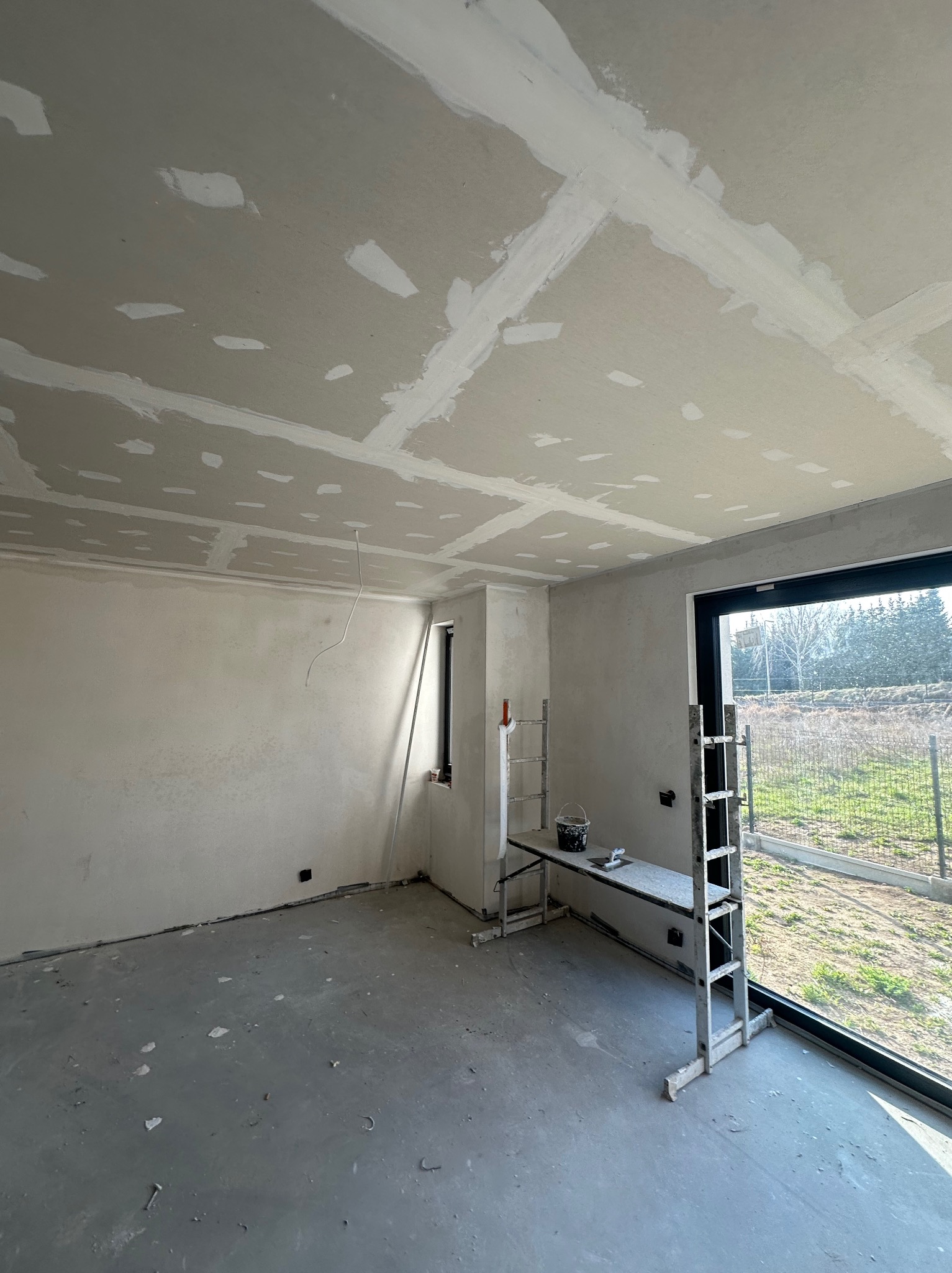 Wnętrze w trakcie prac wykończeniowych: sufit i ściany z płyt gipsowo-kartonowych, widoczne łączenia i szpachlowanie, drabina z narzędziami w pomieszczeniu z oknem.