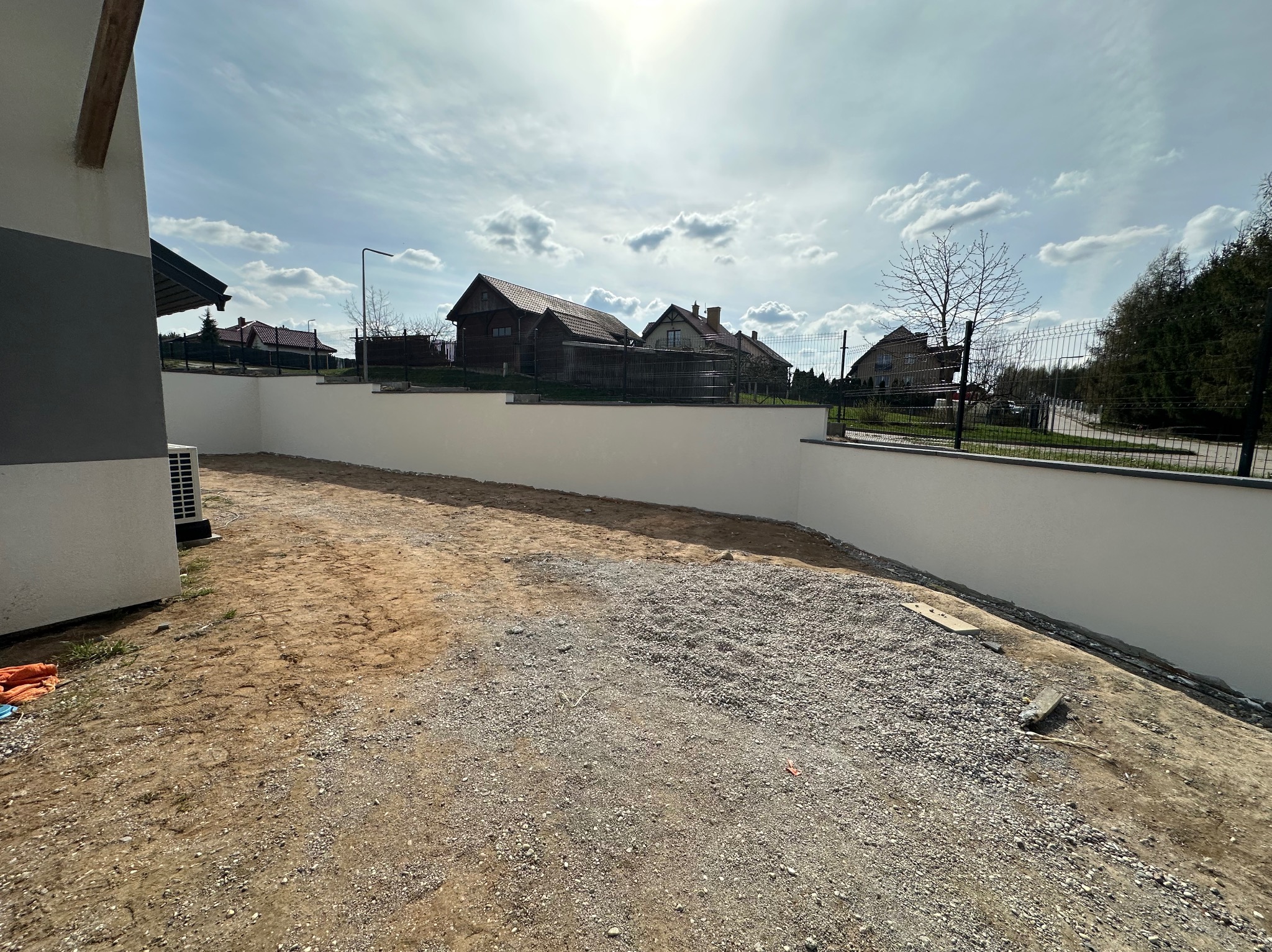 Świeżo pomalowany, biały mur oporowy w ogrodzie, z widocznymi domami w tle i błękitnym niebem z chmurami. Teren przygotowany pod dalsze prace.