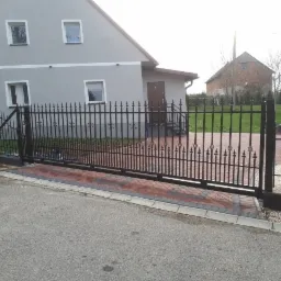 Nowoczesna, przesuwna brama wjazdowa z kutymi elementami dekoracyjnymi, zamontowana przed domem z szarą elewacją, widoczny fragment ogrodzenia i podjazdu z kostki brukowej.