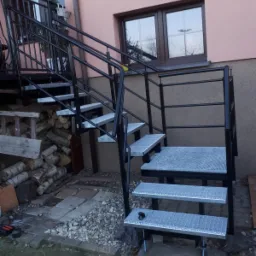 Zewnętrzne schody metalowe z czarną balustradą i ocynkowanymi stopniami, prowadzące do drzwi na piętrze, obok stos drewna i narzędzia.
