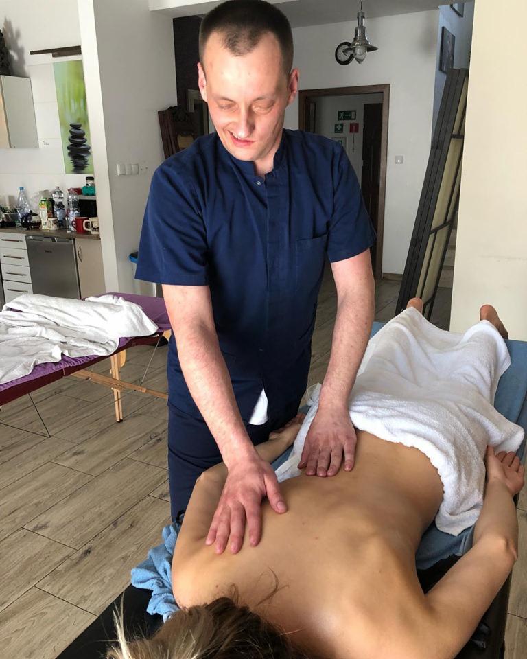 Osoba w granatowym uniformie wykonuje masaż pleców pacjentki leżącej na stole do masażu, widoczny fragment głowy pacjentki, w tle jasne pomieszczenie z wyposażeniem.