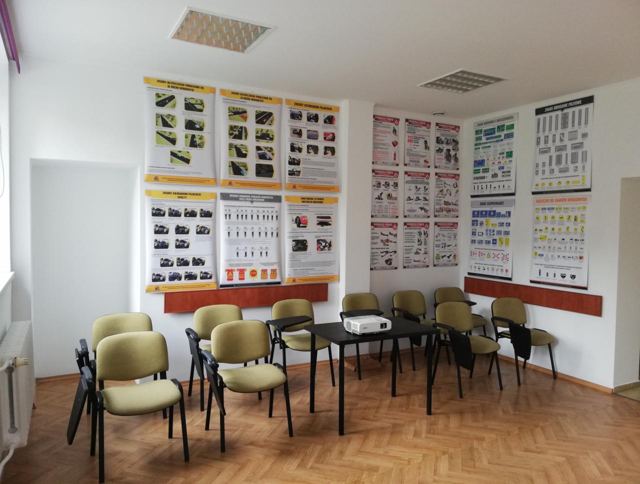 Sala wykładowa z krzesłami, stołem, projektorem i plakatami edukacyjnymi na ścianach, dotyczącymi zasad bezpieczeństwa i znaków drogowych.