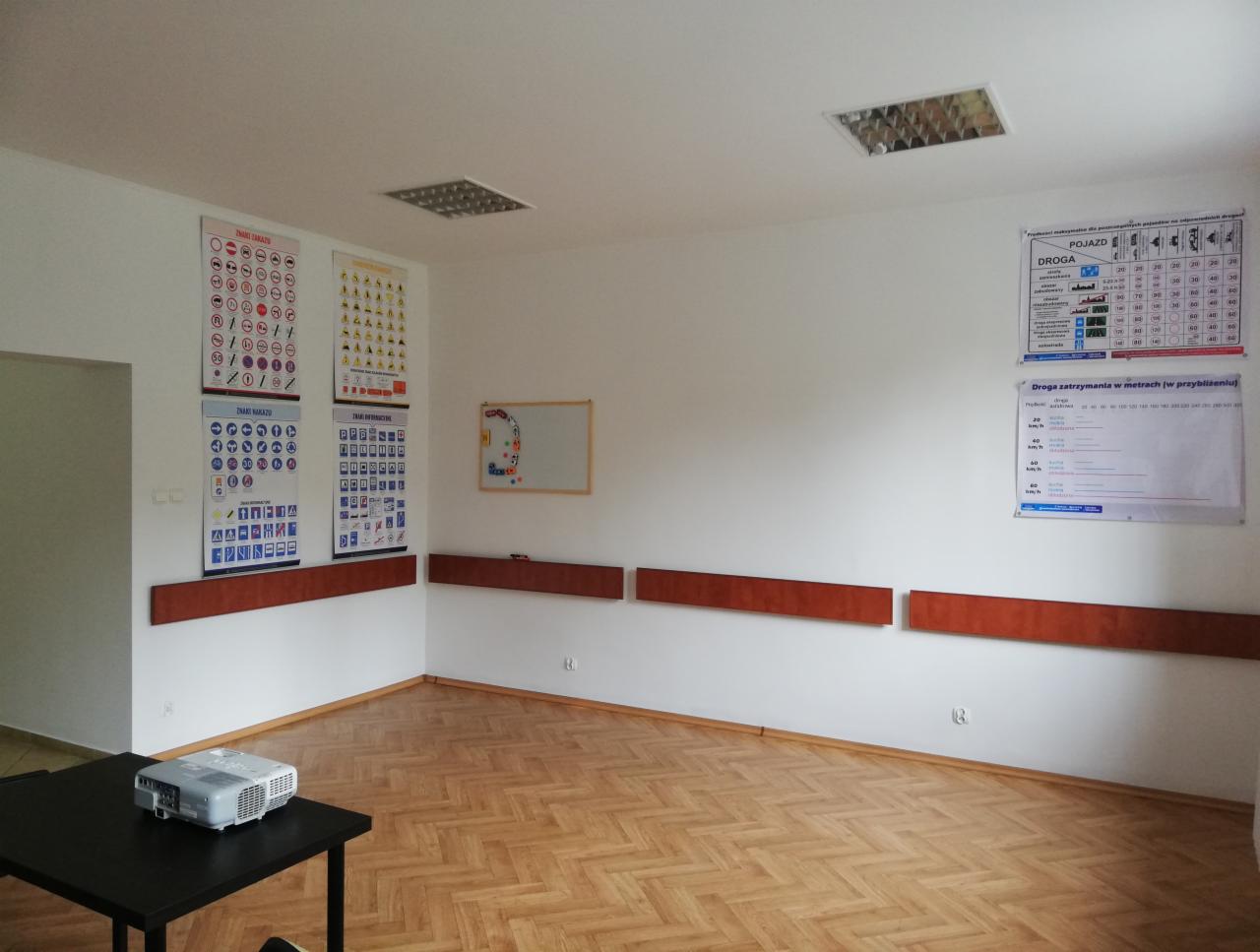 Sala wykładowa z plakatami ze znakami drogowymi na ścianach, projektor na stole, parkiet na podłodze.