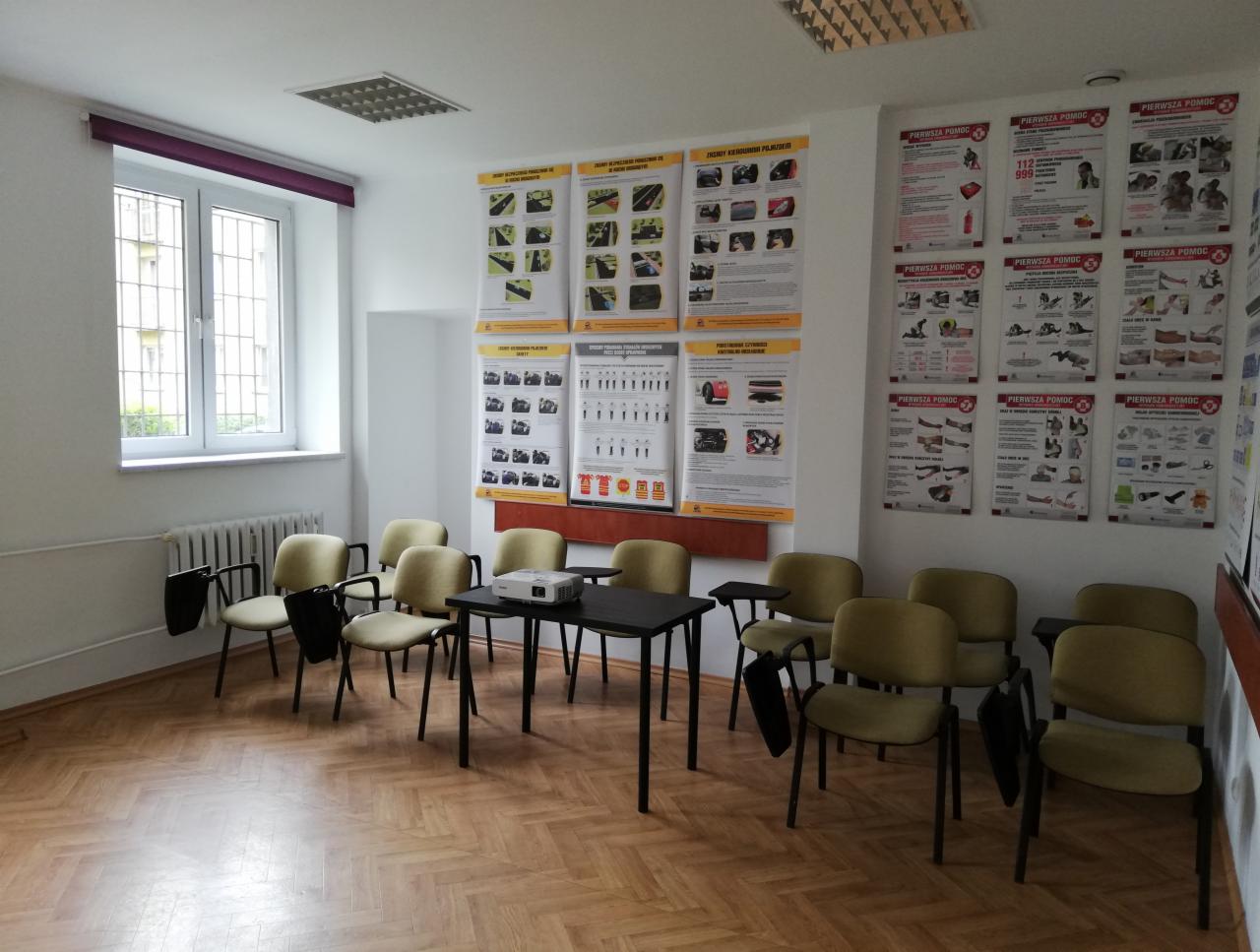Sala szkoleniowa z krzesłami, stołem i projektorem, ściany ozdobione plakatami z instrukcjami pierwszej pomocy, parkiet na podłodze.