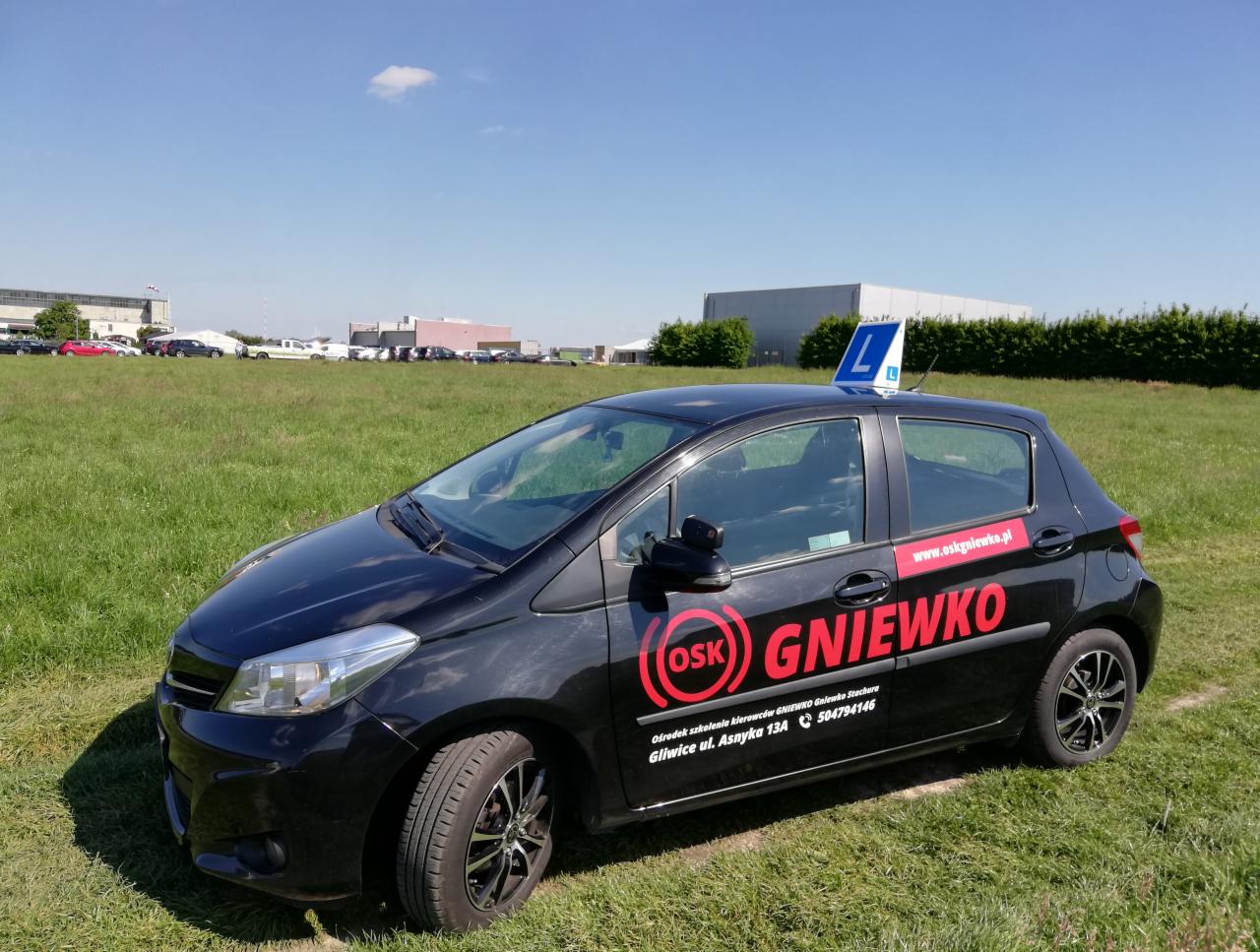 Czarny samochód z oznaczeniem nauki jazdy 'L' na dachu, logo 'OSK GNIEWKO' na boku, stojący na trawiastym terenie z budynkami w tle.