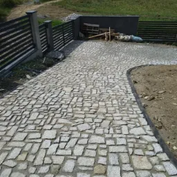 Ułożona z nieregularnych, szarych kamieni kostka brukowa na podjeździe, obrzeże z czarnym krawężnikiem, w tle zielone wzgórze i fragment ogrodzenia.