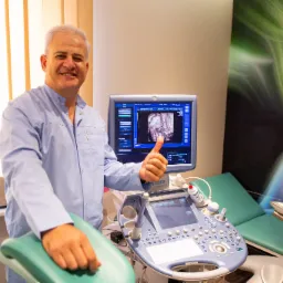 Uśmiechnięty ginekolog w niebieskim uniformie, z uniesionym kciukiem na tle aparatu USG z widocznym obrazem płodu i zielonego fotela ginekologicznego.