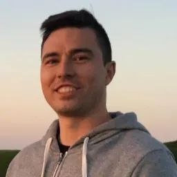 Portret uśmiechniętego mężczyzny w szarej bluzie na tle zachodzącego słońca w Pasłęku. Widok na wzgórza w oddali.