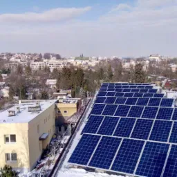 Rząd paneli słonecznych na dachu budynku w zimowej scenerii, z widokiem na miasto w tle.