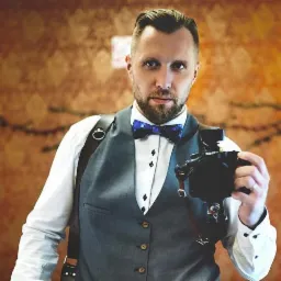 Portret fotografa w eleganckiej kamizelce i muszce w gwiazdy, trzymającego aparat na skórzanych szelkach na tle wzorzystej ściany.