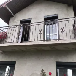 Balkon z metalową balustradą o dekoracyjnych elementach kwiatowych, widok z dołu na elewację budynku z oknami i roletami zewnętrznymi.