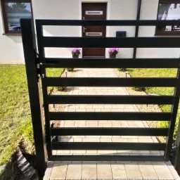 Nowoczesna, czarna furtka z poziomych profili zamontowana w Wieruszowie, widok na ścieżkę z kostki prowadzącą do drzwi wejściowych domu.